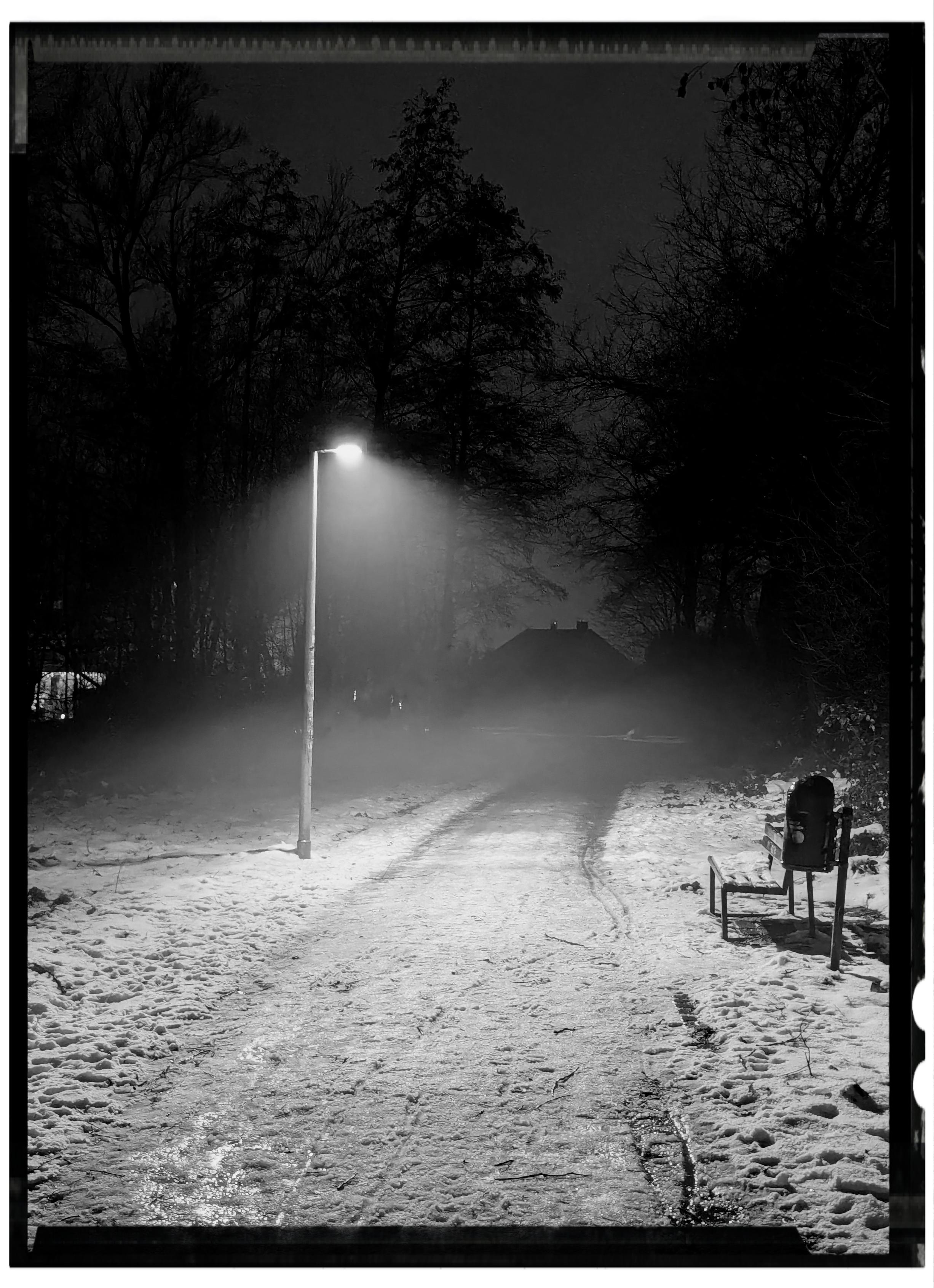 The image is a black-and-white photograph taken outdoors at night.

In the center of the image is a narrow path covered with snow. The path runs from the bottom of the image toward the background. There are visible tracks in the snow along the path.
A single streetlamp stands slightly left of center along the path. The lamp is on and casts light downward, illuminating a circular area of the snowy ground beneath it.
On the right side of the image in the foreground, there is a bench facing the path. Next to the bench is a cylindrical trash bin. Both appear lightly covered with snow.
On both sides of the path are tall trees without leaves. The trees extend from the foreground into the background and fill much of the left and right sides of the image.
In the background, near the end of the path, there is the outline of a building with a sloped roof, partially obscured by darkness and haze.
The sky is dark, and there are no people, animals, or vehicles visible in the image.